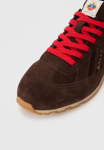 Sapatilhas de camurça castanhas com atacadores vermelhos, marcação dourada, superfície texturizada, bico arredondado e sola de borracha natural.