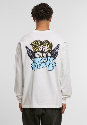 Man with curly hair wearing white long-sleeve shirt featuring two yellow-haired, purple-winged angels and blue stylized text on the back.