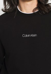 Persona che indossa un maglione nero di Calvin Klein con il logo bianco centrato sul petto e una maglietta bianca visibile al colletto.