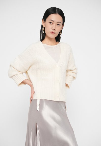 Krém színű kötött kardigán V-nyakkal, gombos záródással és bordázott mandzsettákkal, világos top és oldalhasítékos szatén szoknya fölött viselve.