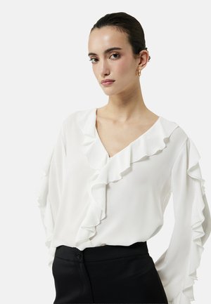 Kvinde med bagstrøget hår iført guldringøreringe, en hvid bluse med flæser og sorte højtaljede bukser mod en ensfarvet baggrund.