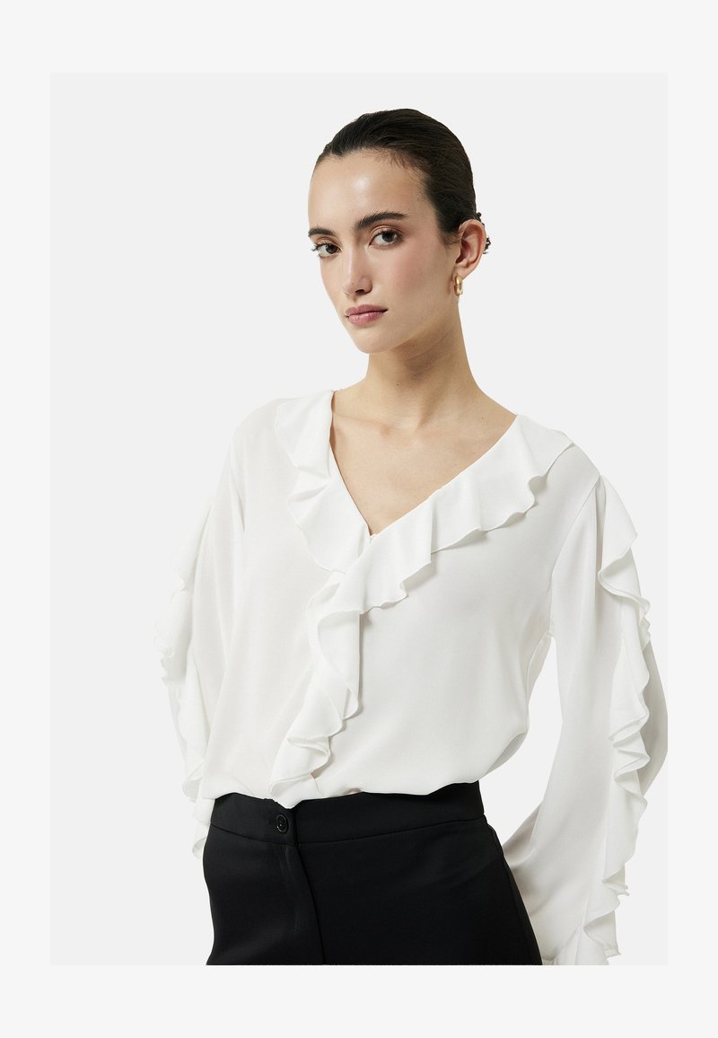 Mujer con el cabello peinado hacia atrás, lleva pendientes de aro dorados, una blusa blanca con volantes y pantalones negros de talle alto sobre un fondo liso.