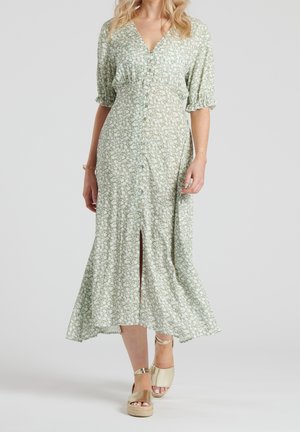 Frau trägt ein hellgrünes, geblümtes Midikleid mit Knöpfen vorne und gerafften Ärmeln bis zum Ellbogen, kombiniert mit goldenen Keilsandalen.