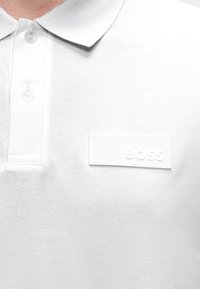 Polo blanc avec col et boutons, avec un patch logo « BOSS » texturé sur la partie gauche de la poitrine.