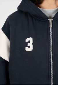 Námornícka mikina s bielym pruhom, zipsovým uzáverom a vyšitým bielym číslom "3" na prednej strane. Mäkká tkanina s voľným strihom.