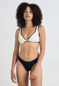 Bikini-setti mustasta ja beigestä, jossa on teksturoitu, uritettu muotoilu, V-pääntie bralette kultaisilla yksityiskohdilla ja matala vyötärö bikinialushousut.