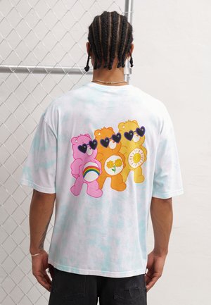 Valkoinen batik-värjätty puuvillainen t-paita, jossa on kolme sarjakuvakarhua eloisissa väreissä; pinkki, oranssi ja keltainen, jotka käyttävät sydämenmuotoisia aurinkolaseja ja pitävät symboleja.