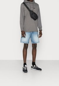 Sudadera gris con capucha, pantalones cortos de mezclilla y zapatillas negras con detalles blancos. El hombre lleva una bandolera negra para un aspecto deportivo.