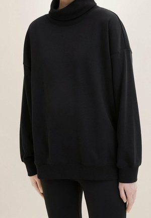 Henkilö, joka on pukeutunut löysään mustaan poolopaitaan, jossa on pitkät hihat ja resorireunukset, yhdistetty mustiin housuihin.