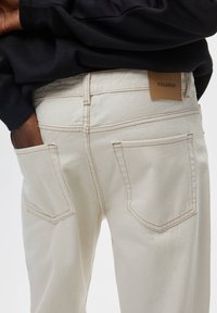 Beżowe spodnie dżinsowe z dwoma tylnymi kieszeniami, z kontrastowymi przeszyciami i brązową skórzaną metką "PULL&BEAR" nad prawą kieszenią.