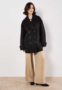 Cappotto doppiopetto nero realizzato in misto lana con grandi bottoni, colletto a risvolto e maniche ampie, abbinato a pantaloni beige a gamba larga.