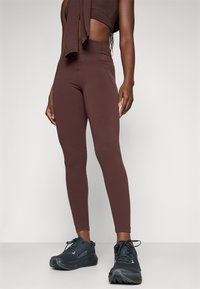 Mujer vistiendo leggings marrones de cintura alta y zapatillas de correr negras, de pie contra un fondo claro y liso.