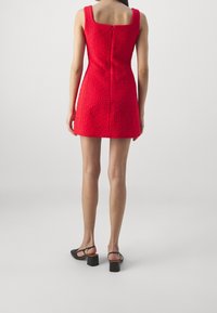 Punainen lyhyt mekko, jossa on neliönmuotoinen kaula-aukko ja teksturoitu kangas. Varustettu takana olevalla vetoketjulla ja istuvalla siluetilla, yhdistetty mustiin korkokenkiin.