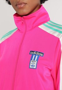 Primer plano de una persona que lleva una chaqueta de entrenamiento Adidas de color rosa brillante con franjas en azul y blanco y un parche del logo de Adidas en el pecho.