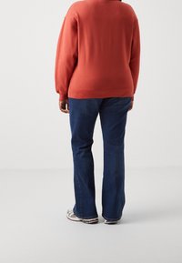 Personne portant un sweat à capuche orange brûlé, un jean bleu et des baskets blanches, se tenant de dos contre un fond blanc uni.