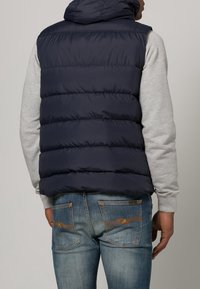 Mann, der eine marineblaue Steppweste über einem hellgrauen Hoodie und blauen Jeans trägt, von hinten vor einem schlichten Hintergrund betrachtet.
