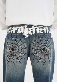 Baksidan av blå jeans med svart spindelnätsbroderi och små röda spindlar på varje ficka, i kombination med en vit skjorta.