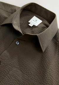 Camisa marrón texturizada con cuello estructurado, cierre de botones y un bolsillo de parche. La etiqueta indica "NEXT", corte regular, talla M.