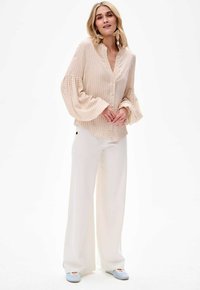 Bluzka w jasnobeżowe paski z kołnierzem mandarynki i rozszerzanymi rękawami, w połączeniu z białymi spodniami o szerokich nogawkach i jasnoniebieskimi butami.