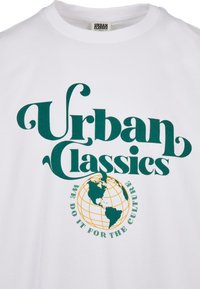 Fehér pamut póló, zöld és sárga grafikus nyomattal, amelyen "Urban Classics" felirat és földgömb dizájn látható, ívelt betűtípussal kiemelve.