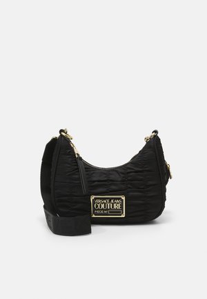 Sac d'épaule matelassé noir avec une large bandoulière ajustable et une plaque dorée indiquant Versace Jeans Couture Piece No.