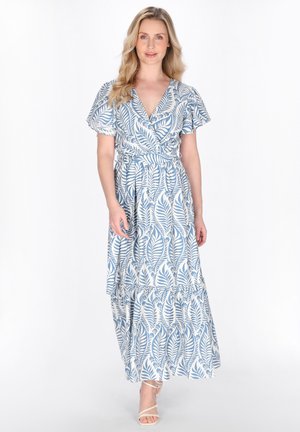 Kvinde stående iført en blå og hvid slå-om-kjole med bladmønster, flagrende ærmer og beige hæle med stropper mod en hvid baggrund.