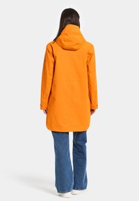 Ljusorange vattentät jacka med avslappnad passform, huva och långa ärmar, kombinerad med vida blå jeans och vita sneakers.