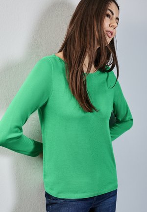 Groene lange mouwen trui gemaakt van zachte stof, met een ronde hals en een getailleerde pasvorm. Draagt met blauwe spijkerbroek.