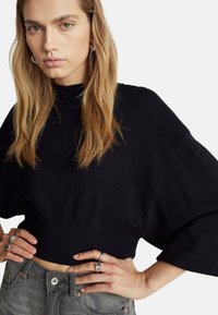 Černý zkrácený svetr s vysokým výstřihem a širokými rukávy. Model má na sobě šedé džíny a stříbrné prsteny, charakterizovaný jemnou, texturovanou látkou.