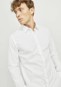 Camisa blanca de manga larga con cuello abotonado, que presenta un sutil patrón texturizado y botones claros, de ajuste entallado.