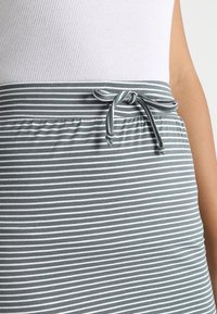 Zone de taille d'une personne portant un top côtelé blanc et une jupe grise avec des rayures horizontales blanches et un nœud attaché à la taille.