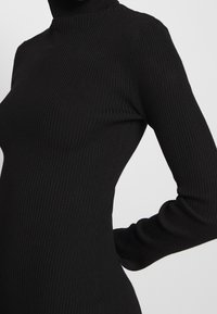 Närbild av en person som bär en svart ribbad långärmad polokrage tröja med armen böjd bakom ryggen.