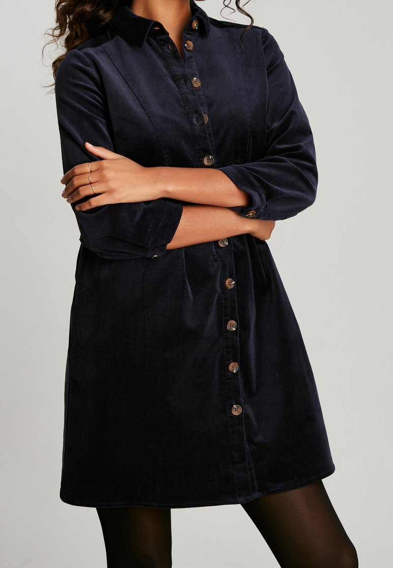 Granatowa sukienka koszulowa z korduroyu z zapięciem na guziki, kołnierzykiem oraz podwiniętymi rękawami. Posiada kieszenie z przodu i lekko rozszerzany dół.