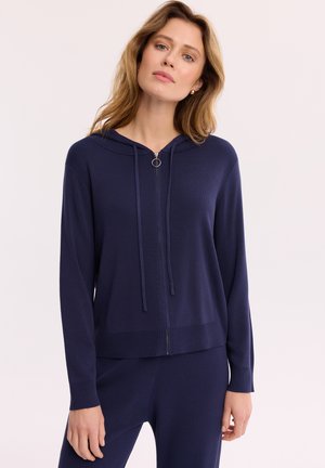 Kobieta ubrana w granatową bluzę z kapturem na zamek błyskawiczny i sznurki oraz pasujące spodnie, stojąca na tle jednolitego jasnego tła.