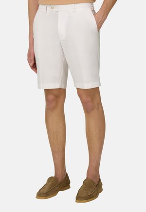 Mann trägt weiße kniehohe, maßgeschneiderte Shorts mit Seitentaschen und braune Wildleder-Slipper, steht vor einem einfarbigen Hintergrund.