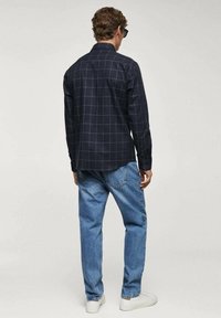Uomo che indossa una camicia a maniche lunghe a scacchi blu navy, jeans blu, sneaker bianche e occhiali da sole, in piedi rivolto di spalle su uno sfondo semplice.