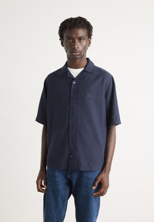 Marineblauw overhemd met korte mouwen en knoopsluiting, voorzien van een omklapbare kraag en een vierkante patchdetail. Gecombineerd met denim jeans, met een ontspannen pasvorm.