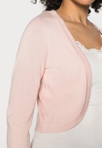 Lichtroze gebreide cardigan met een cropped ontwerp, open voorkant en lange mouwen, gecombineerd met een witte geribde tanktop eronder.