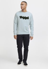 Felpa azzurra con testo decorativo nero "Original" al centro, abbinata a pantaloni neri e sneakers nere.