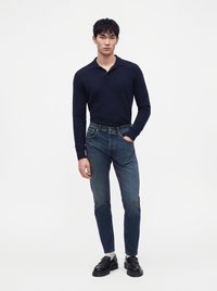 Jeune homme debout, portant un polo à manches longues sombre, un jean bleu, des chaussettes blanches et des mocassins noirs, sur un fond uni clair.