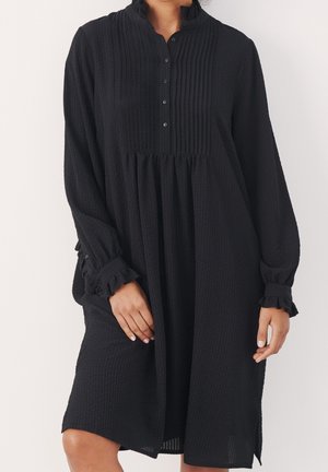 Frau trägt ein schwarzes, strukturiertes langärmeliges Kleid mit geknöpftem Kragen und Rüschen an den Manschetten, steht vor einem weißen Hintergrund.