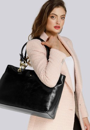 Donna con blazer rosa pallido strutturato che tiene una grande borsa in pelle nera sulla spalla, sfondo neutro.