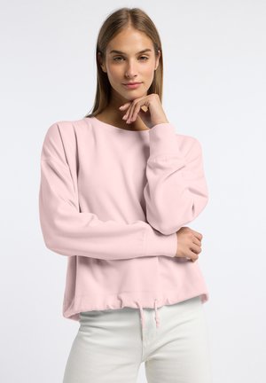 Młoda kobieta z prostymi blond włosami nosi jasnoróżową bluzę z długim rękawem i białe spodnie, pozując z ręką blisko twarzy.