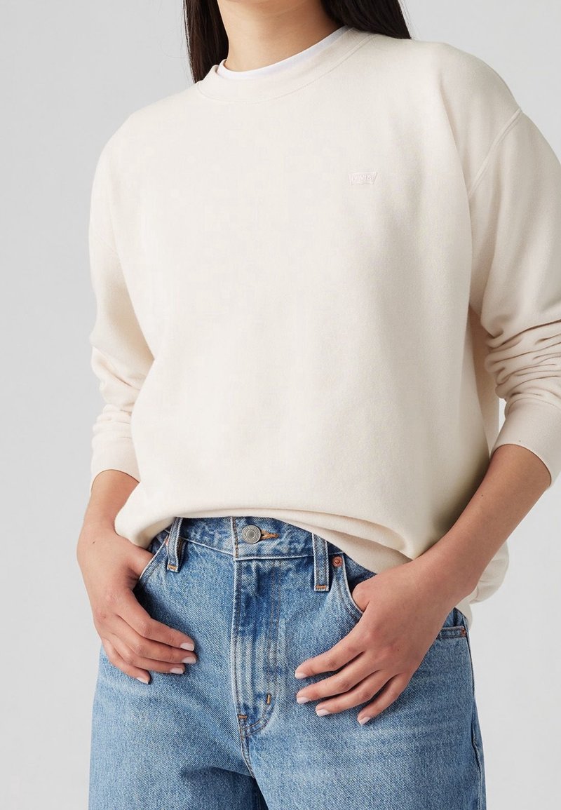 Personne portant un sweat-shirt crème à col rond, rentré dans un jean bleu clair taille haute, les mains reposant près des poches avant.
