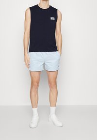 Marinblå ärmlös topp med vit logotyp, kombinerad med ljusblå shorts. Modellen har på sig vita sneakers och synliga vita ankelsockor. Enkelt design.
