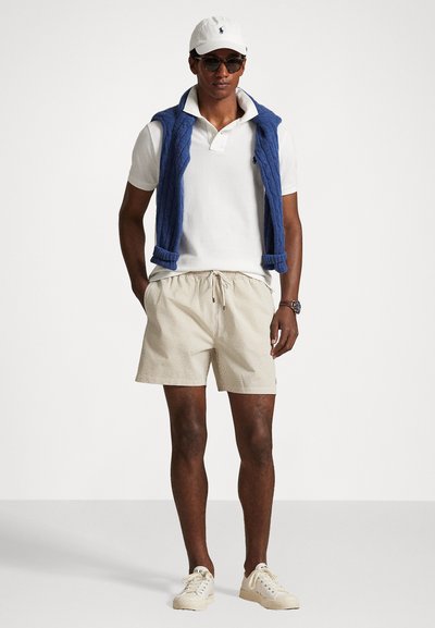 Πολυμερής μπλούζα και ριγέ σορτσάκι, με έναν μπλε γιλέκο από πάνω. Τα αξεσουάρ περιλαμβάνουν λευκό καπέλο και γυαλιά ηλίου. Φωτεινές αθλητικές παπούτσια ολοκληρώνουν το σύνολο.