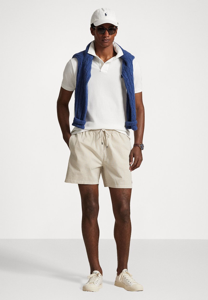 Polohemd und gestreifte Shorts, kombiniert mit einem blauen Strickweste. Die Accessoires umfassen eine weiße Kappe und Sonnenbrillen. Helle Sneakers runden das Outfit ab.