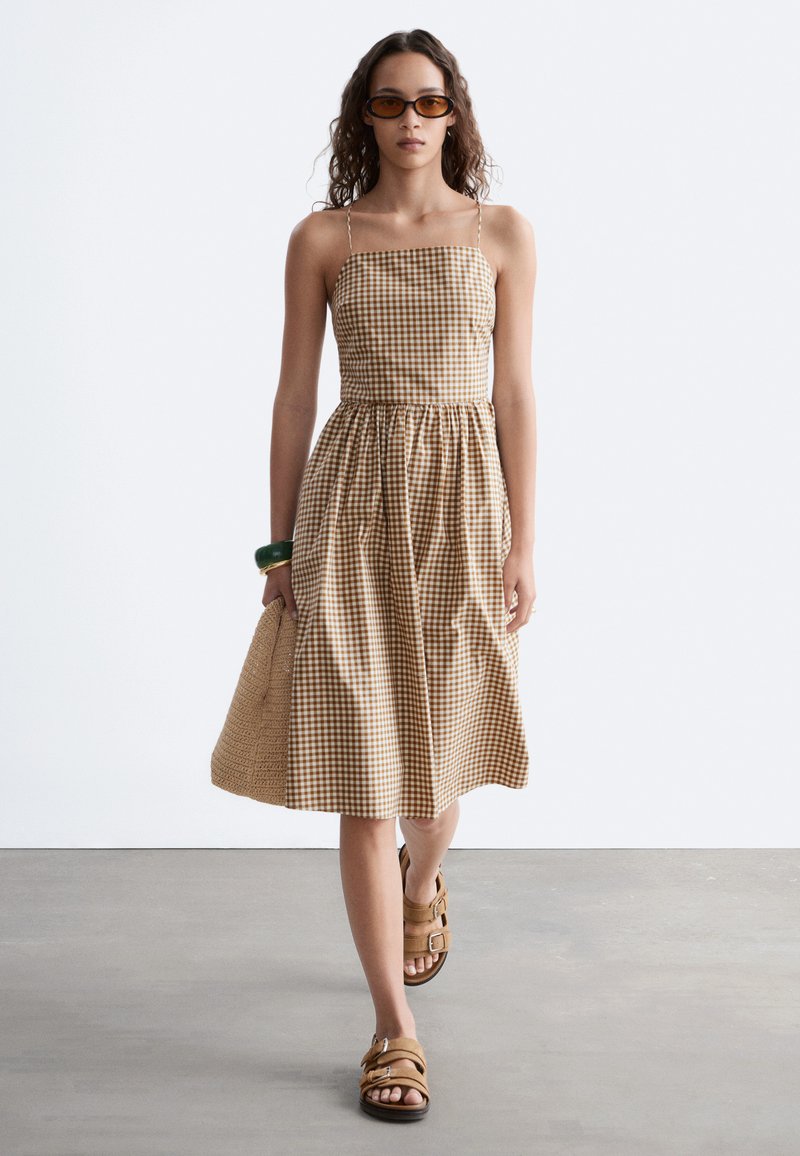 Jeune femme marchant portant une robe sans manches à carreaux marron et blanc, des sandales marron, des lunettes de soleil orange, un bracelet vert, et portant un sac tressé.