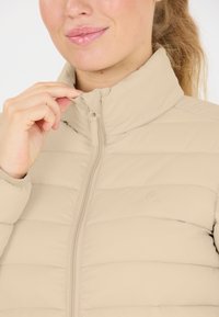 Beige puffer jakke med quiltet tekstur og opretstående krave. Har en frontlynlås og logo detalje, der fremviser et glat, blødt stof.