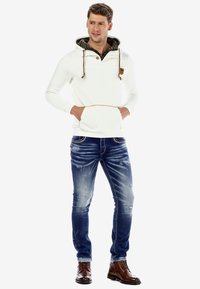 White pullover hoodie with drawstrings, button, and front pocket; paired with distressed blue jeans and brown lace-up boots.
